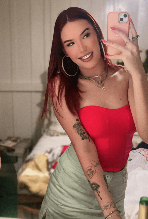 Young woman posing in a mirror selfie wearing a pink strapless top and light green skirt, with tattoos on her left arm and large hoop earrings, holding a pink phone.