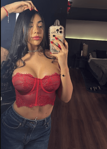 Woman taking a mirror selfie wearing a red lace bustier and jeans, in a dimly lit room with dark wood floor.