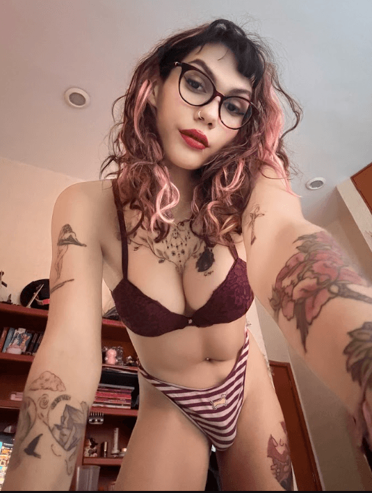 Self-portrait of a person with pink-tinted hair and glasses wearing burgundy lace bra and striped underwear in a bedroom, showing tattoos on arms and chest.